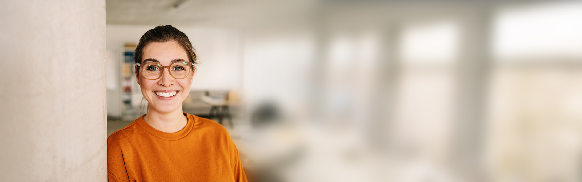 eine Frau in einem orangefarbenen Hemd und einer Brille, die an eine Wand gelehnt ist