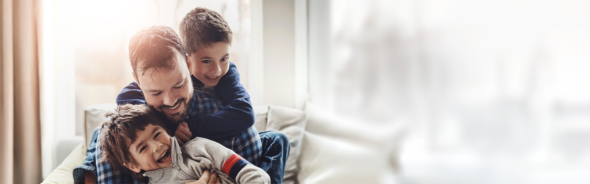 Alleinerziehender Vater sitzt im Wohnzimmer und umarmt lachend seinen einen Sohn, während der zweite Sohn ihn von hinten umarmt.
