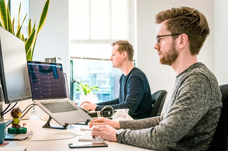 Zwei Männer in Pullovern sitzen an nebeneinanderstehenden Schreibtischen vor ihren Laptops und Bildschirmen.